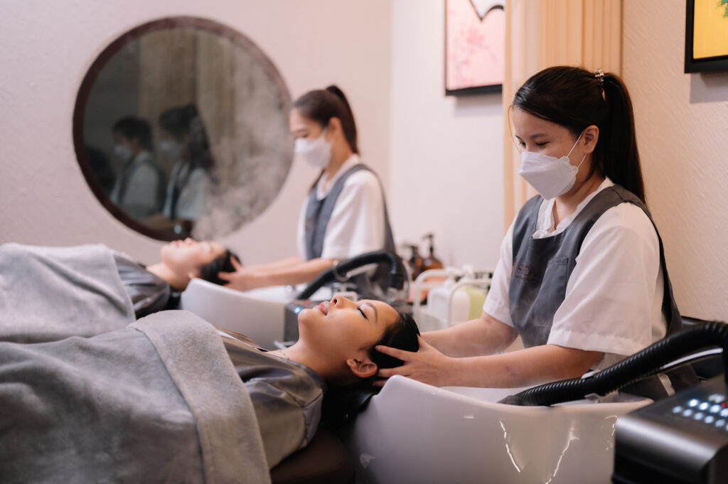 Therapists at Siesta Head Spa and Salon in Bangkok providing relaxing head spa and scalp care treatments. Two clients are receiving gentle scalp massages at a comfortable shampoo station, with steam rising for an enhanced spa experience. The serene spa environment promotes relaxation and rejuvenation, featuring professional scalp massage services to improve scalp health and hair wellness.